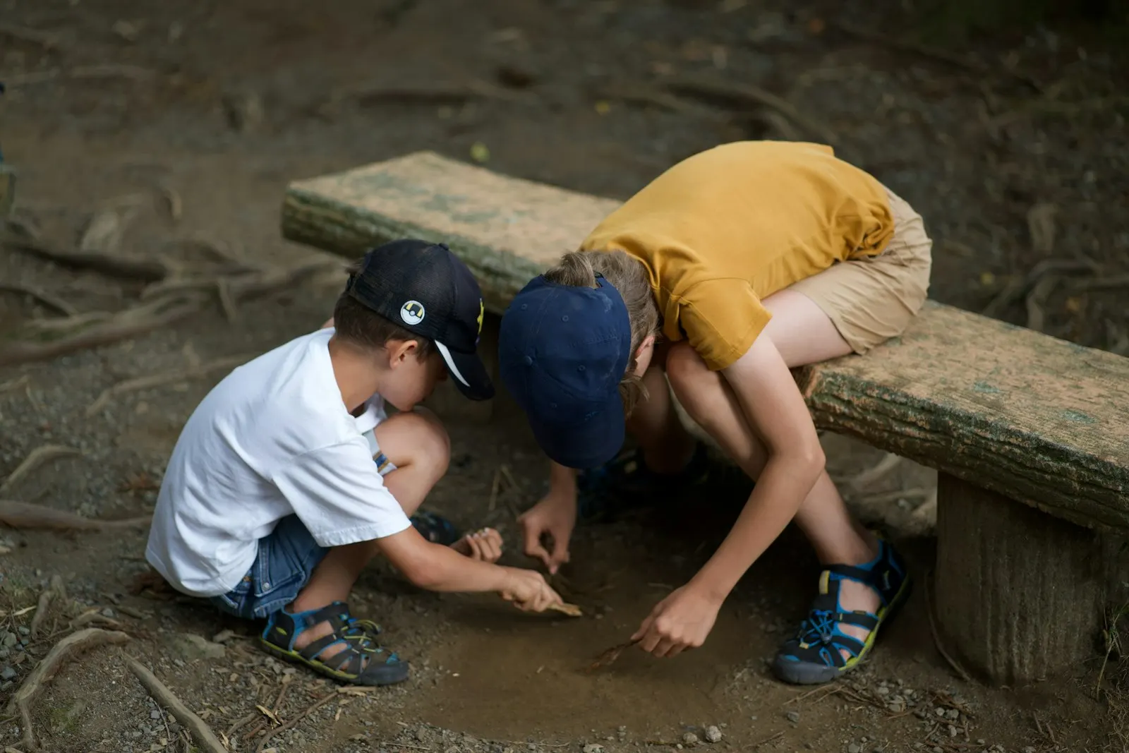 Dos niños agachados buscando algo en el suelo, debajo de un banco
