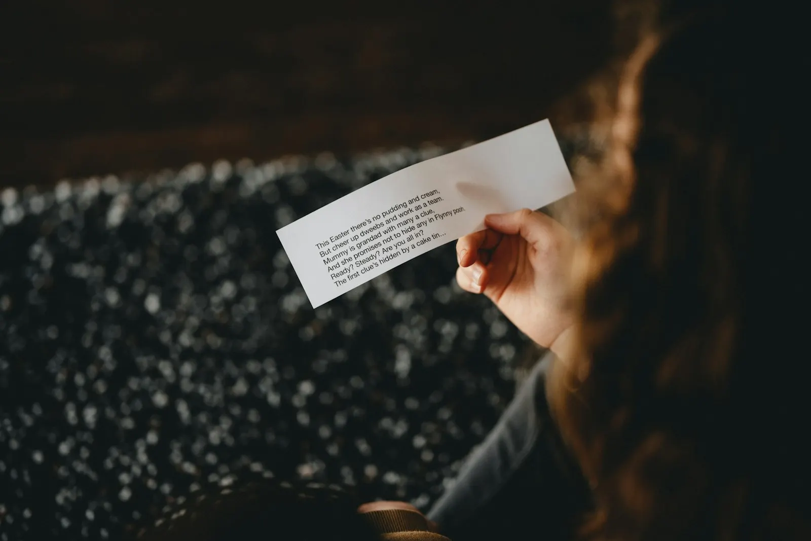 Una mano sostiene una nota de papel pequeña con un texto escrito, en una habitación en penumbra